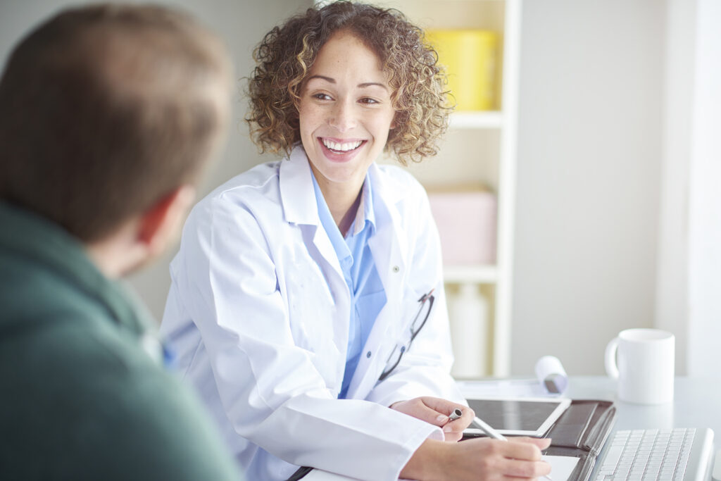 A smiling female hearing care provider in a white coat engages in a friendly conversation with a patient in an office, holding a pen while taking notes. A tablet, keyboard, and coffee mug are visible on the desk, suggesting a professional setting.
