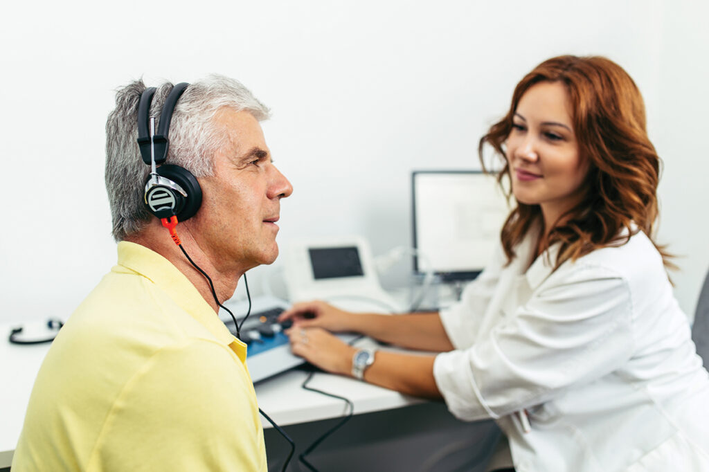A senior man wearing headphones undergoes a professional hearing test with a smiling hearing care professional in a clinical setting. The hearing care professional is adjusting the equipment while monitoring the test results on a computer screen.