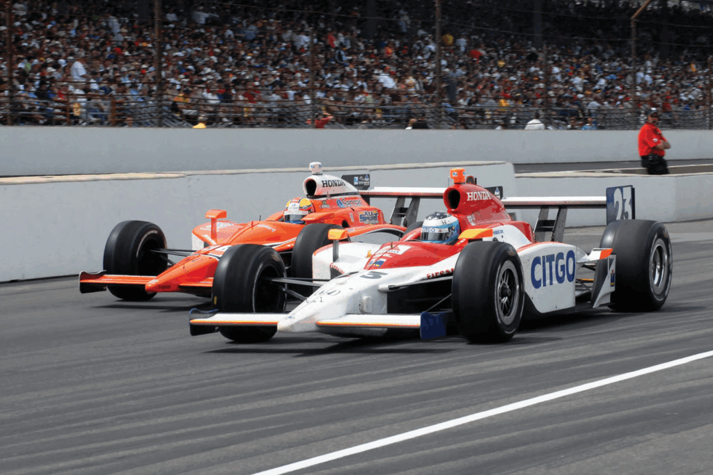 Two IndyCars racing side-by-side at the Indy 500 with a packed crowd in the stands