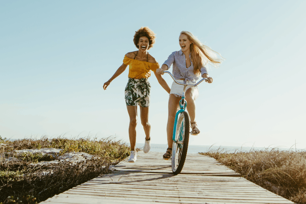 Two women laughing and enjoying a sunny day — one riding a bicycle and the other running beside her along a boardwalk by the ocean.