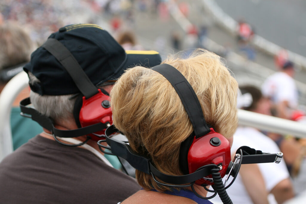 A couple wearing red over-ear hearing protection headsets at a NASCAR race, seated in the grandstands with the track and crowd in the background.