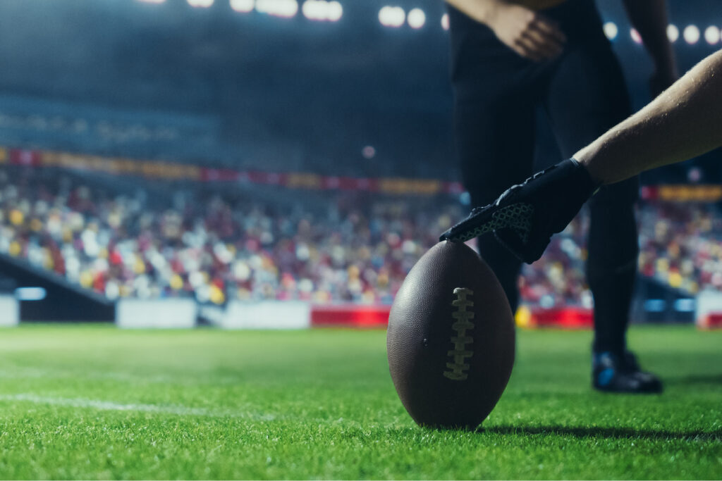 Close-up of a football player’s hand in a glove holding an American football on the field, preparing for kickoff in a stadium.