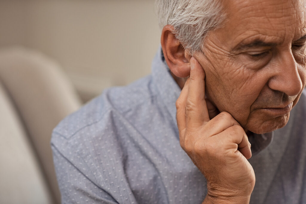 Close-up of a senior man sitting indoors, touching his ear with a worried expression, suggesting possible hearing difficulty.