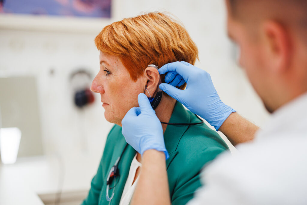 Hearing care professional fitting a hearing aid on a woman with short red hair during an appointment. The provider is wearing blue gloves while adjusting the real ear measurement device behind her ear.