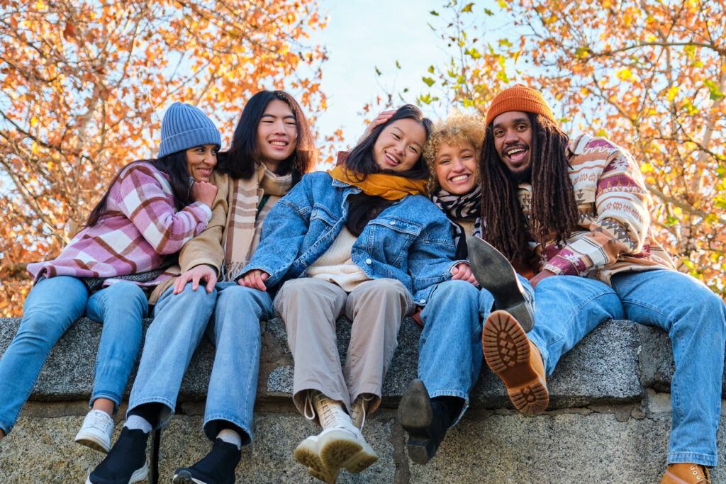 Group of young adults in fall clothing sitting on a stone wall, smiling and laughing together with colorful autumn leaves in the background.