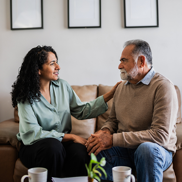 A younger woman offers reassurance by gently placing her hand on the shoulder of an older man as they sit together in a comfortable living room setting.