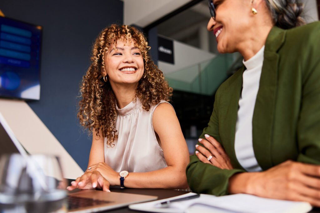 Two professional women smile and chat across a desk in an office setting, suggesting friendly collaboration and supportive conversation.