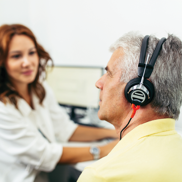 An man wears headphones during a professional hearing test while his hearing care provider monitors the assessment on a computer nearby.