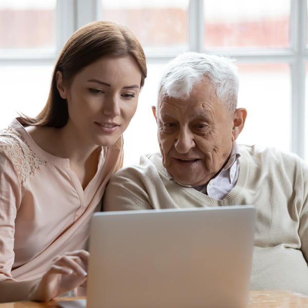 A young woman helps an older man look at information on a laptop at home. They appear focused and engaged, exploring hearing health resources together online.