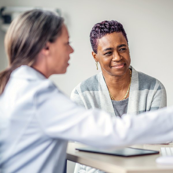 A middle-aged woman smiles while talking with her hearing care provider during a hearing consultation. The provider gestures toward a tablet as they discuss test results in a bright, welcoming clinic setting.
