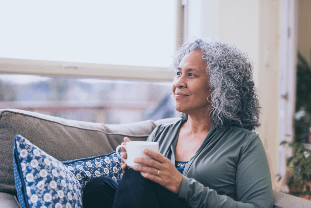 A woman with gray curly hair relaxes on her couch with a warm drink, looking out the window peacefully. She appears content and reflective, representing the confidence and clarity that come from better hearing health.