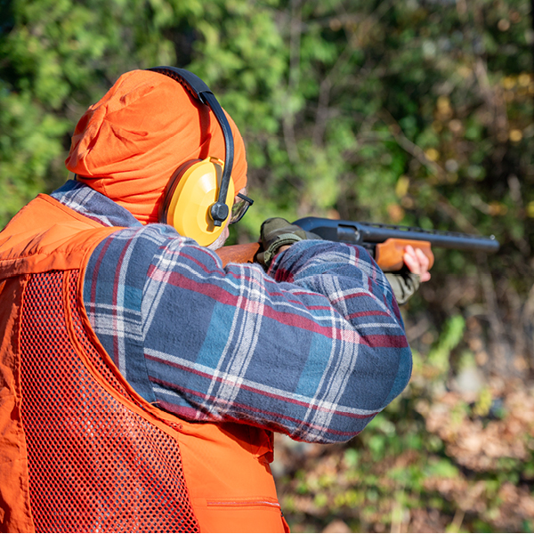 Hunter aiming a shotgun while wearing orange hearing protection earmuffs and safety gear in a wooded area.