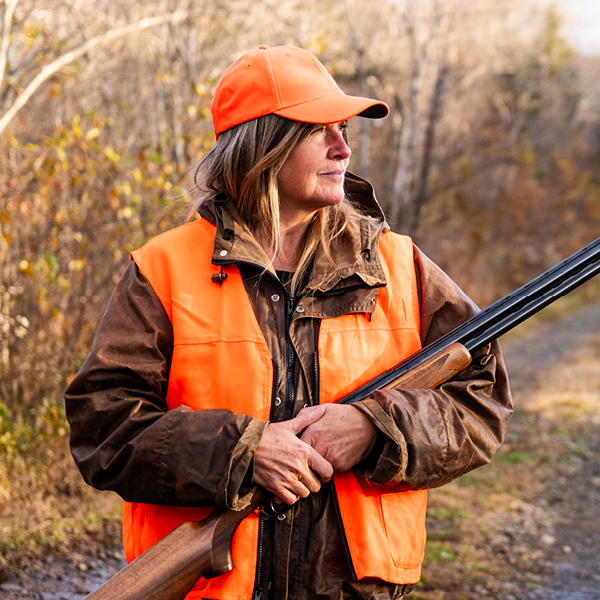 Female hunter wearing an orange cap and vest, holding a shotgun outdoors during fall hunting season.