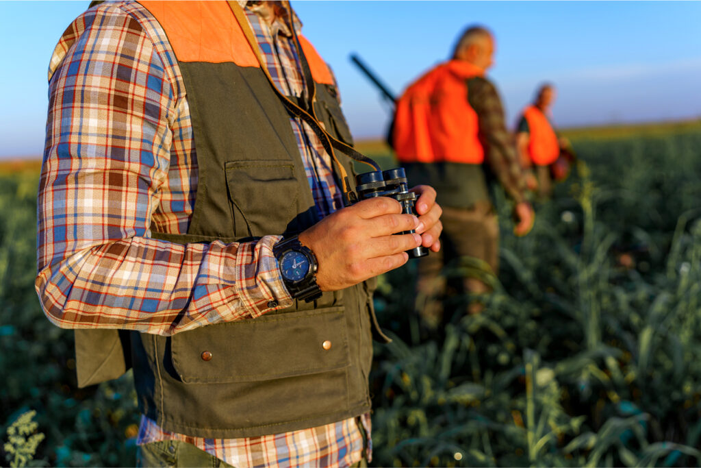 Close-up of a hunter wearing a plaid shirt and safety vest, holding binoculars in the field alongside other hunters in orange vests.