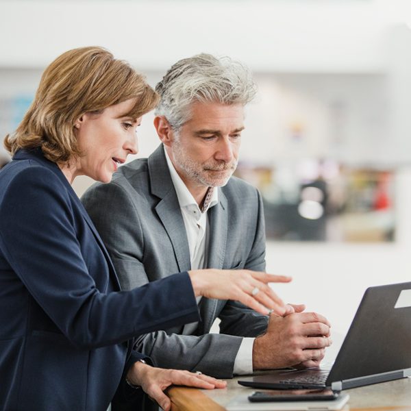 A professional woman gestures toward a laptop screen while discussing something with a man in a business setting. Both appear focused and engaged.