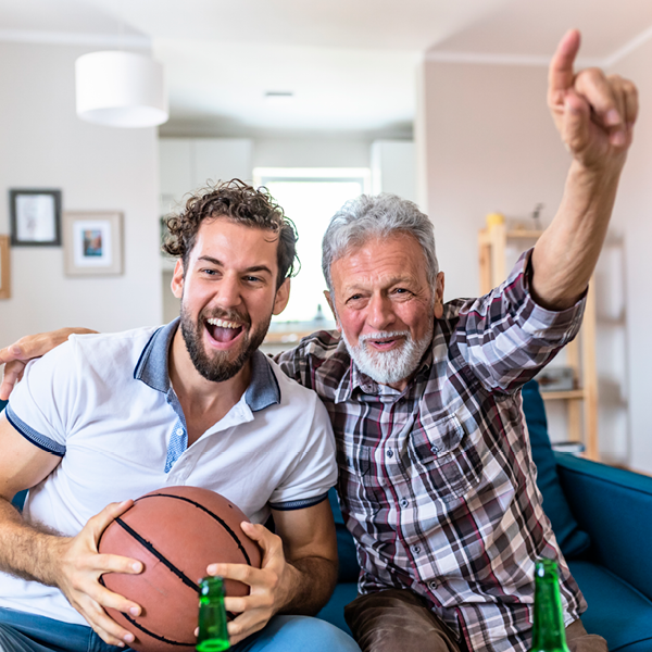 Smiling father and adult son watching a basketball game together at home, cheering and celebrating a big play while enjoying the action on TV.