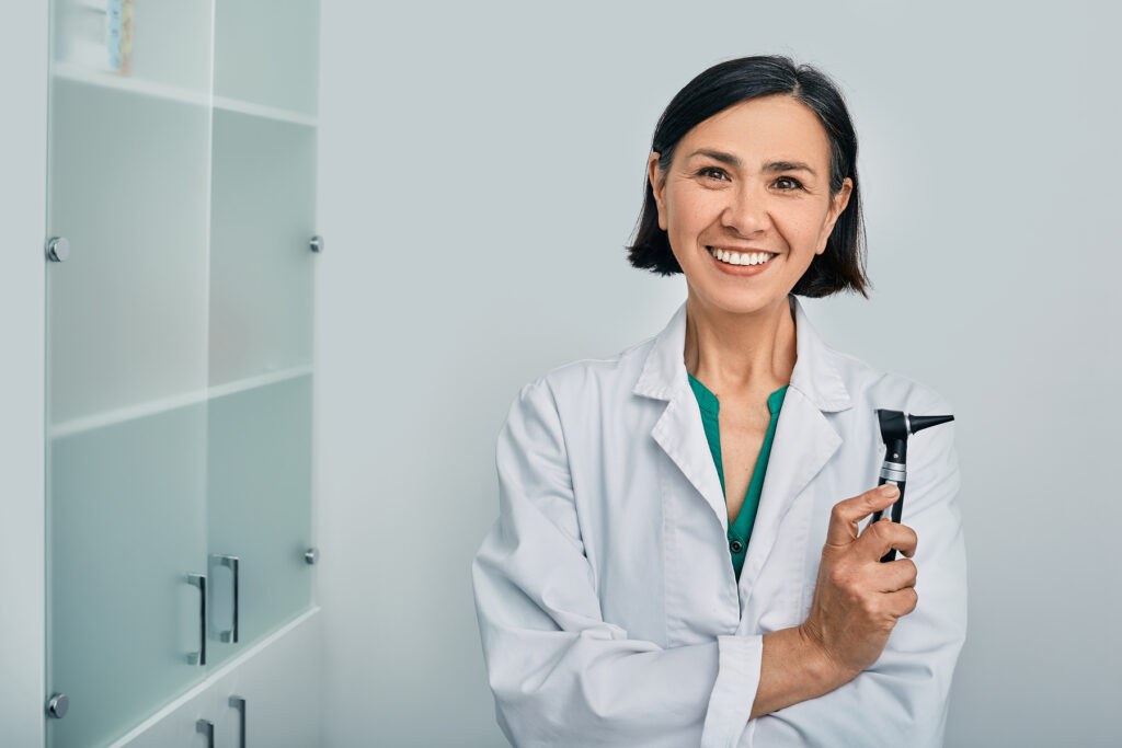 A confident audiologist holding an otoscope smiles in her clinic, representing trusted professional hearing care.