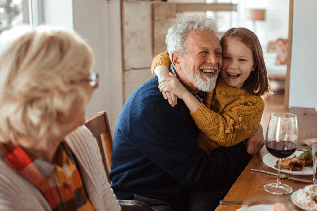 Grandfather joyfully hugging his granddaughter at the dinner table while family shares a meal, symbolizing connection and the importance of hearing health.