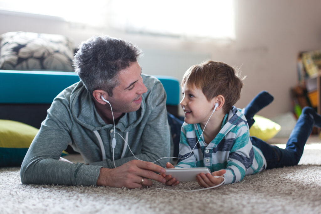Father and young son lying on the floor sharing earbuds and listening to audio together.