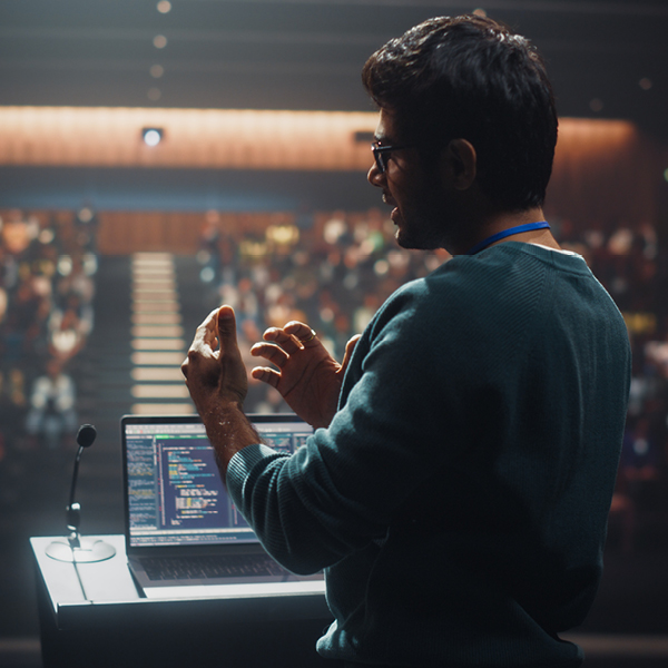 Man speaking on stage in front of an audience with a laptop open beside him during a presentation.
