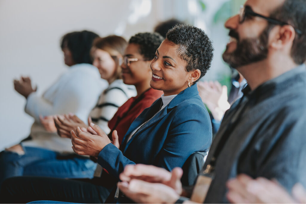 Group of people sitting in rows and smiling while applauding during an indoor event or presentation.