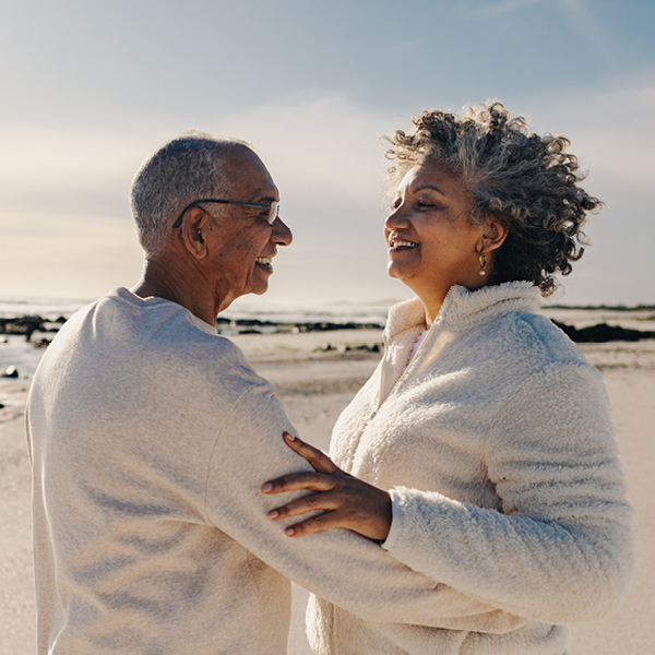 An aging couple smiling and holding each other while standing on a sunny beach. The man is wearing a hearing aid.