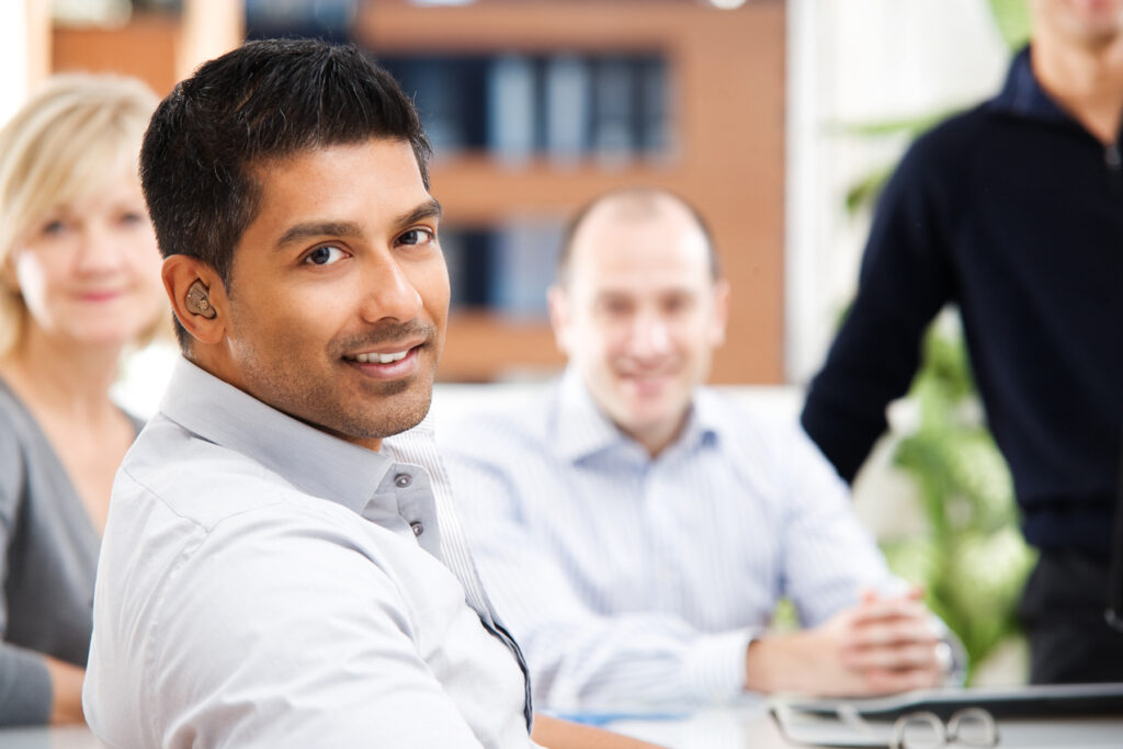 Man wearing discreet hearing aids smiling during a meeting with colleagues in a bright office.