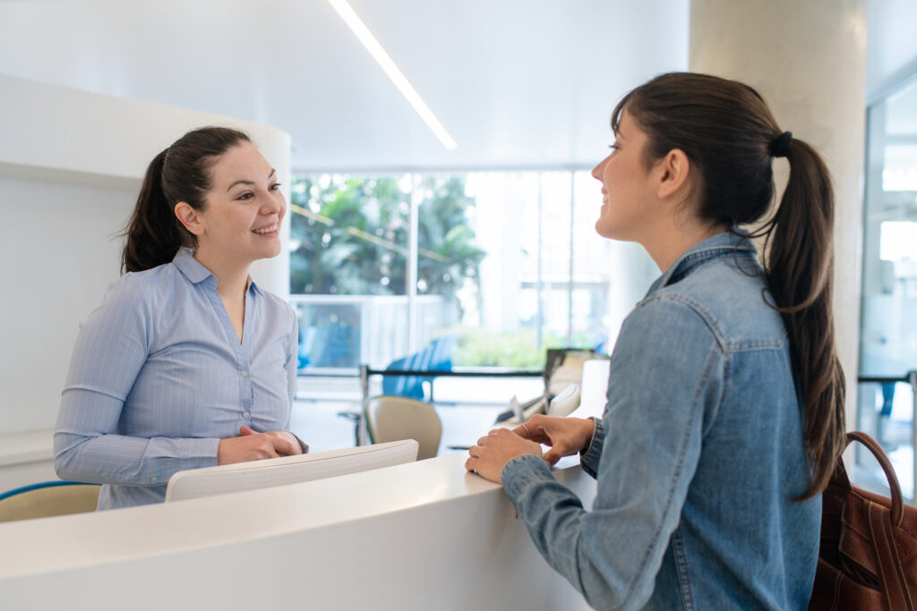 A woman checks in at a hearing clinic reception desk while speaking with a friendly front-office staff member.