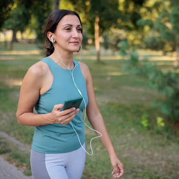 Woman walking in a park while listening to music through earphones, illustrating active living and mindful hearing care for brain wellness.