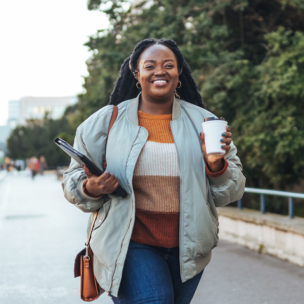 Woman walking confidently with a coffee cup and tablet, symbolizing focus, independence, and the benefits of protecting brain and hearing health.