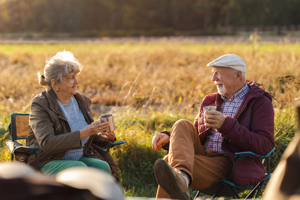 Aging couple sitting outdoors enjoying coffee together on a sunny day, smiling and talking — representing connection, communication, and healthy aging with hearing wellness.