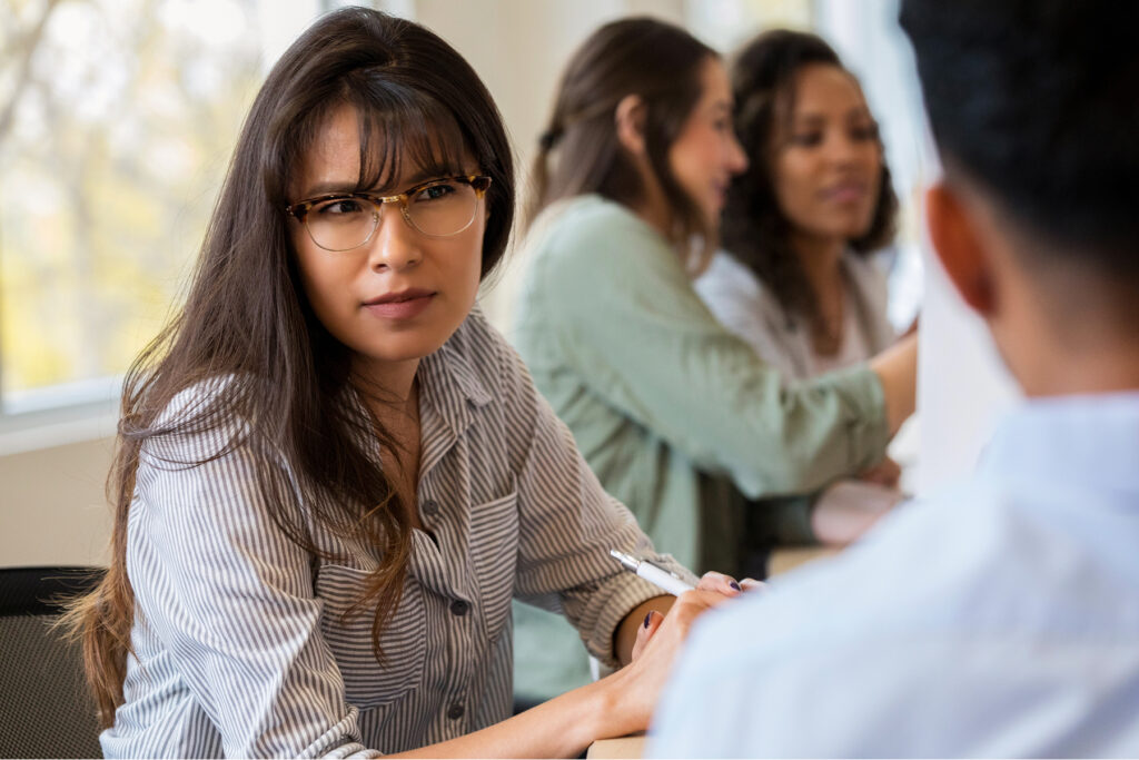A woman in a meeting leans forward with a confused expression, appearing to strain to understand someone speaking.