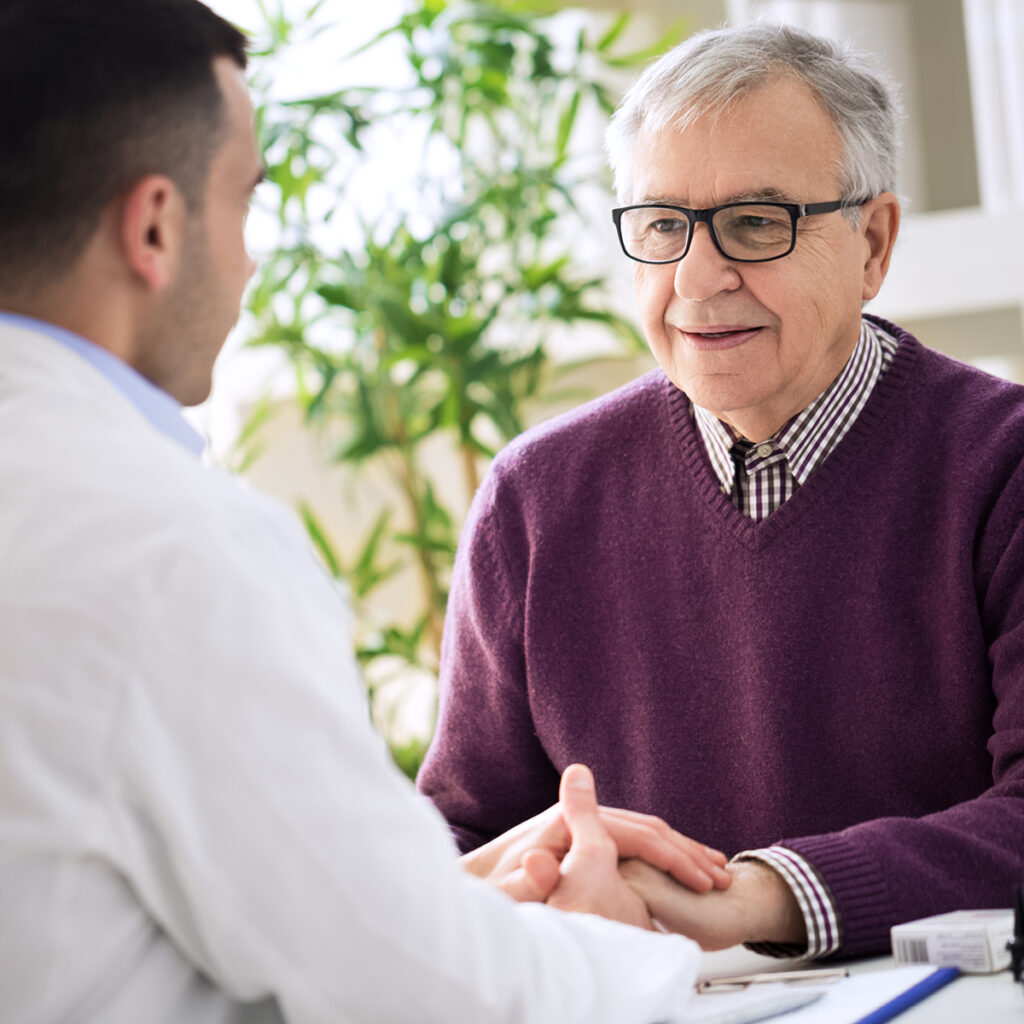 An older man meets with his audiologist, holding hands in a reassuring, attentive hearing care consultation.
