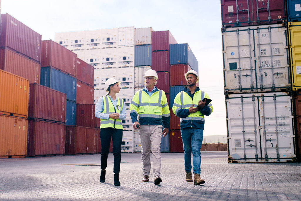 Dockworkers and supervisors wearing high-visibility safety gear walk through a shipping container yard at an active port.