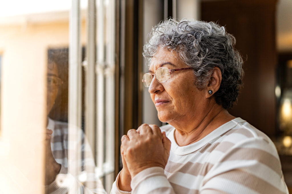 Older woman reflecting by a window, representing social isolation and cognitive changes associated with untreated hearing loss and dementia.