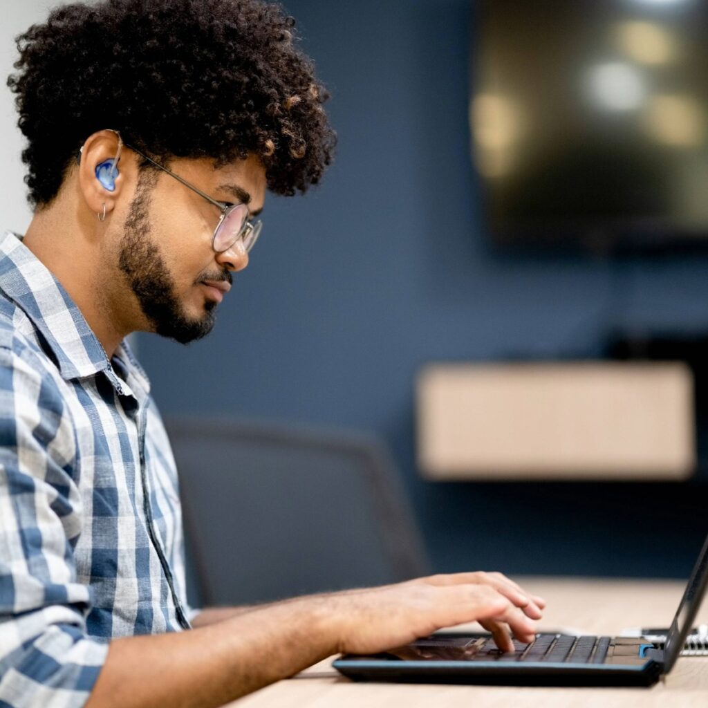 A young man wearing hearing aids works on a laptop during a focused listening and communication task.
