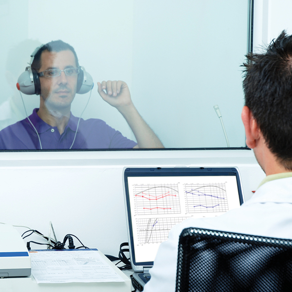 Adult patient wearing headphones during a professional hearing test with a hearing care professional reviewing results on a computer.