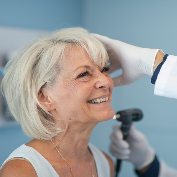 Older woman smiling during a professional hearing exam as a clinician examines her ear in a clinic.