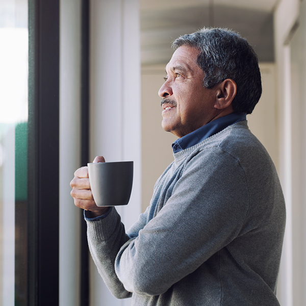 Older man holding a coffee cup while looking out a window, symbolizing dual sensory health and aging-related vision and hearing loss.