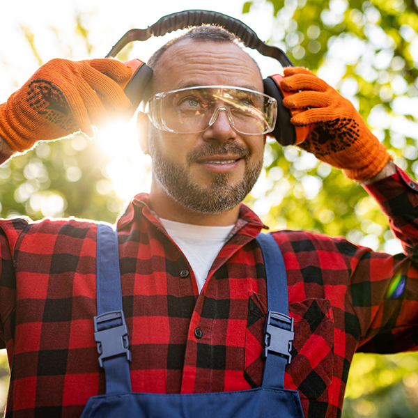 Farmer in red flannel shirt and safety goggles adjusting over-ear hearing protection before operating loud farm machinery, emphasizing year-round hearing safety in agriculture.