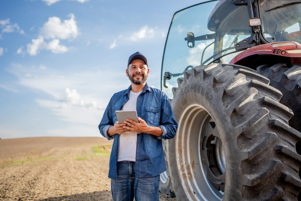 Farmer standing beside a large tractor tire in an open field, holding a tablet, highlighting agricultural work environments where prolonged farm noise can increase risk of hearing loss.