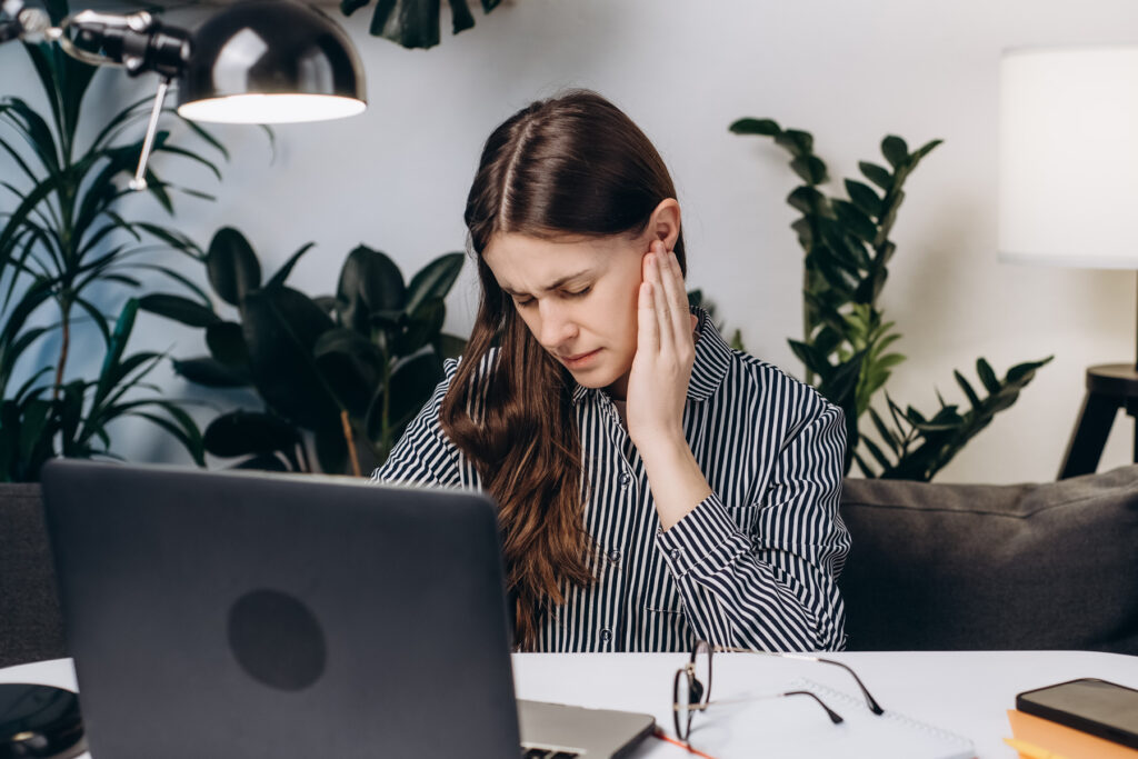 Woman sitting at a laptop holding her ear in discomfort, illustrating tinnitus symptoms such as ringing or buzzing interfering with daily work.