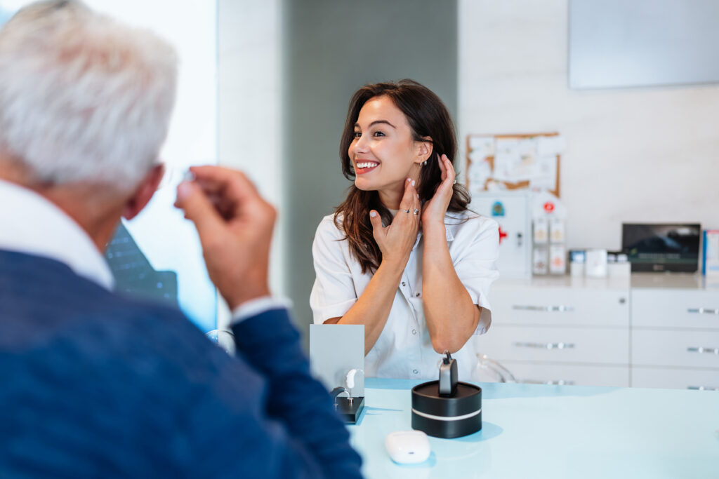 Audiologist smiling while adjusting a patient’s hearing aid during a certified hearing care appointment in a modern clinic.