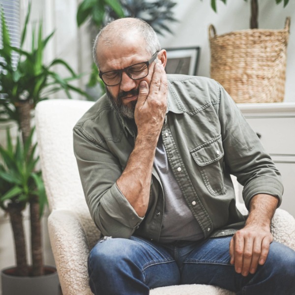 Man sitting in chair holding his jaw and ear in discomfort, illustrating how tinnitus or ear-related symptoms can cause distress and require professional hearing evaluation.