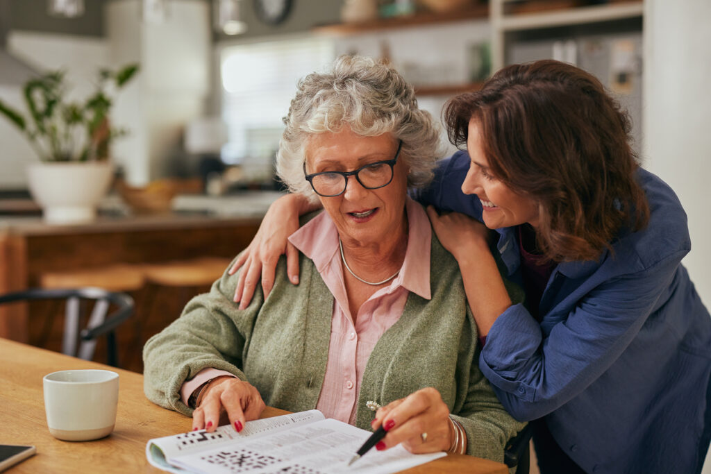 Adult daughter helping an older woman complete a crossword puzzle at a kitchen table, symbolizing cognitive engagement and brain health.