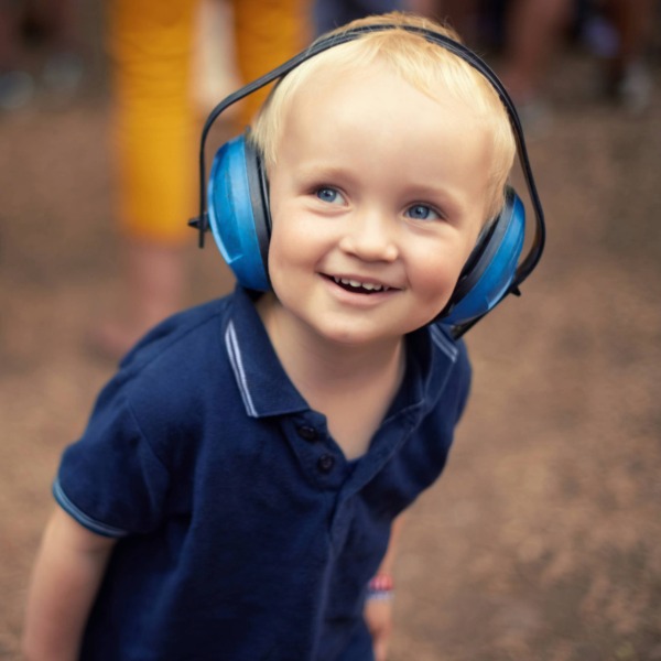 A young toddler with blonde hair and blue eyes smiles broadly while wearing blue protective ear muffs and a navy polo shirt, standing outdoors at an air show.
