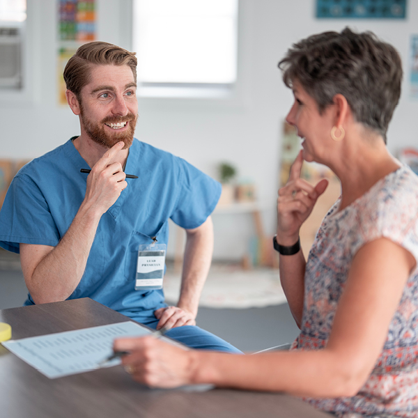 A male healthcare provider in blue scrubs smiles warmly while consulting with a middle-aged woman seated across a table, with paperwork in front of them in a bright clinical setting.