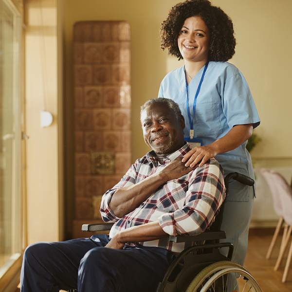 A female healthcare provider in light blue scrubs stands behind an older adult man seated in a wheelchair, smiling as she rests a hand on his shoulder in a warm, softly lit care facility.
