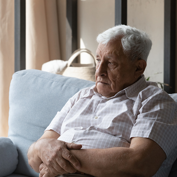 An older man sits alone on a couch with arms crossed, looking away with a distant expression