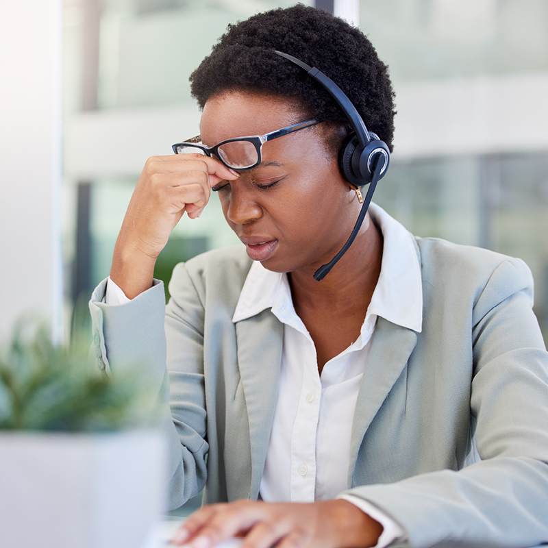Woman wearing a headset and glasses pinching the bridge of her nose in frustration while working at a computer, showing signs of listening fatigue